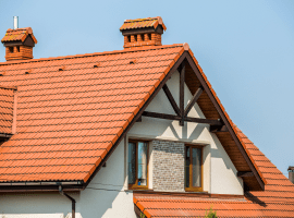 A close-up architectural photograph of a traditional European-style house roof with terracotta orange tiles. The roof features a prominent triangular dormer window with exposed dark brown wooden beams creating a Tudor-style truss design. The dormer has a white-painted brick facade and two matching brick chimneys with orange clay caps. The main roof has a steep pitch and is covered with uniform terracotta tiles in a herringbone pattern. The image is taken against a clear light blue sky. The architectural details include clean lines, precise craftsmanship, and a symmetrical design. The photograph is cropped to focus on the upper portion of the roof and dormer, showing the geometric patterns and architectural elements in sharp detail.