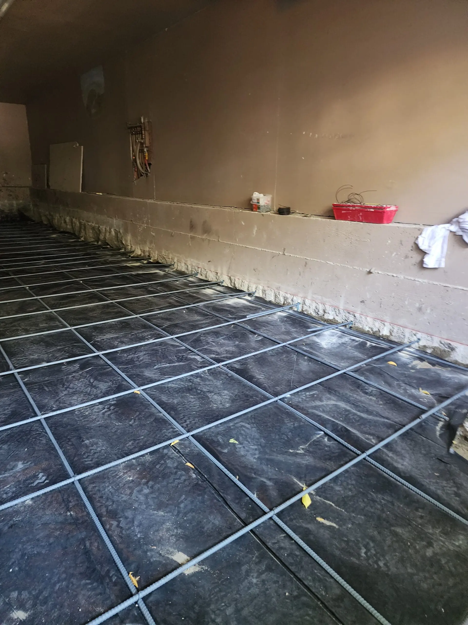 A dimly lit interior room with dark slate-gray floor tiles arranged in a grid pattern. The tiles are rectangular and have a matte finish with subtle variations in color and texture. The floor has visible grout lines between each tile. The walls are painted in a light beige color and show signs of wear and aging, with visible water damage and discoloration near the floor line. A red plastic container sits on the right side of the wall. The lighting is warm and creates shadows in the corners of the room. The ceiling is visible at the top of the frame and shows some electrical fixtures mounted on it. The overall composition has a moody, industrial aesthetic with the contrast between the dark floor and light walls.