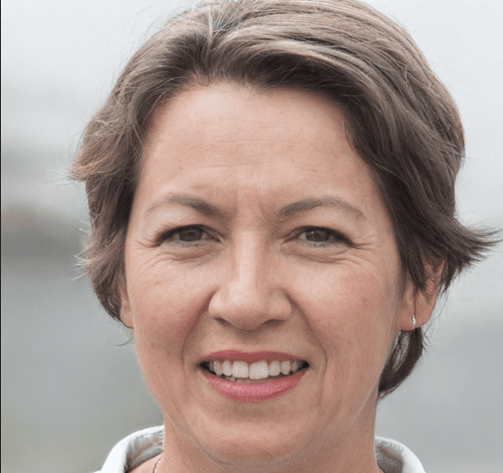 A professional headshot portrait against a soft, blurred background. The subject has short brown hair styled in a casual updo with some strands framing the face. They are wearing a white collared shirt and small silver stud earrings. Their expression shows a warm, genuine smile with bright white teeth visible. The skin tone is light, and there are natural wrinkles around the eyes and mouth from the expression. The lighting is soft and even, creating gentle shadows and highlights on the face. The image has a professional, polished quality with sharp focus on the facial features while maintaining a natural, approachable feel. The background is in muted tones of gray and white, creating a clean, uncluttered composition.