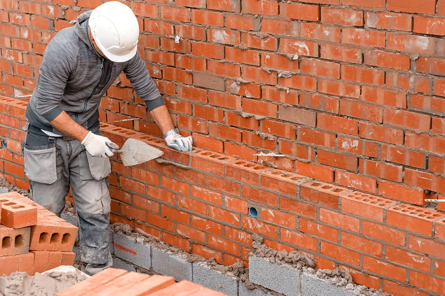 A construction worker in a gray long-sleeve shirt and light gray pants with utility pockets, wearing a white hard hat and white work gloves, is laying red clay bricks in a bricklaying project. The worker is bending forward, using a gray trowel to apply mortar between the bricks. The wall being built is partially complete, with gray concrete blocks visible at the base and bottom edge. The existing brick wall is made of standard orange-red bricks in a traditional running bond pattern. The bricks show signs of weathering and age with some discoloration and wear marks. The scene is photographed from a low angle, showing the worker's profile against the brick wall. In the foreground, there are several loose red clay bricks ready for use in the construction. The lighting is natural and even, showing a daytime work setting.