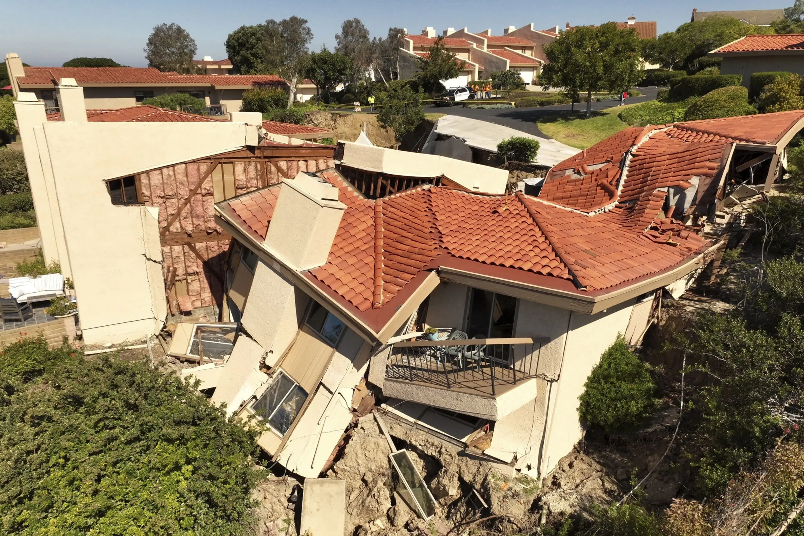 An aerial photograph of a severely damaged residential building with terracotta-colored tile roofing. The two-story structure has white stucco walls and is partially collapsed, with sections of the building tilting dramatically and falling into the ground. The roof tiles are displaced and scattered across the scene. The building features a balcony with metal railings that is partially detached. The surrounding area shows green vegetation and other residential buildings with similar terracotta roofs in the background. The damage is extensive, with structural elements displaced and exposed. The image is taken from a high angle, showing the full scope of the structural failure. The sky is clear and blue, and the scene is in a suburban setting with other houses visible in the background.