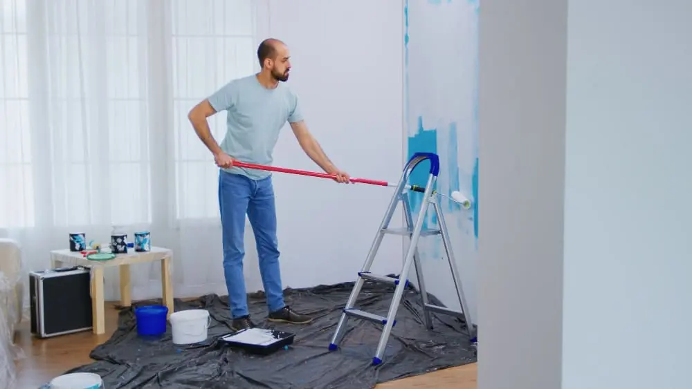 A photograph of a middle-aged man painting a white wall with blue paint using a long-handled roller extension. The person is wearing a light blue t-shirt, blue jeans, and black shoes, standing on a black drop cloth laid out on the floor. A metal step ladder is positioned next to the wall for reaching the higher areas. On the left side of the room, there is a wooden side table with a black speaker and several blue paint cans. A blue bucket sits on the drop cloth near the ladder. The room has white walls and natural lighting coming from a window with sheer white curtains visible in the background. The wall being painted shows blue paint starting from the middle height and extending upward. The image is shot in a bright, well-lit interior space with hardwood flooring partially visible at the bottom of the frame.