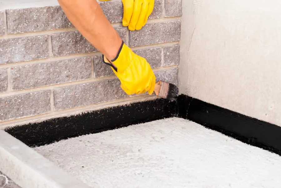 A close-up photograph of a construction worker applying black waterproofing material to a concrete foundation. The worker is wearing bright yellow rubber gloves and is using a brown paintbrush to apply the material along the base of a gray brick wall. The wall is made of uniform light gray bricks with visible mortar lines. The concrete foundation is smooth and white, creating a stark contrast with the black waterproofing material being applied. The image is cropped to focus on the application process, showing only the hands, forearms, and the corner where the wall meets the foundation. The lighting is bright and even, typical of daytime outdoor conditions.