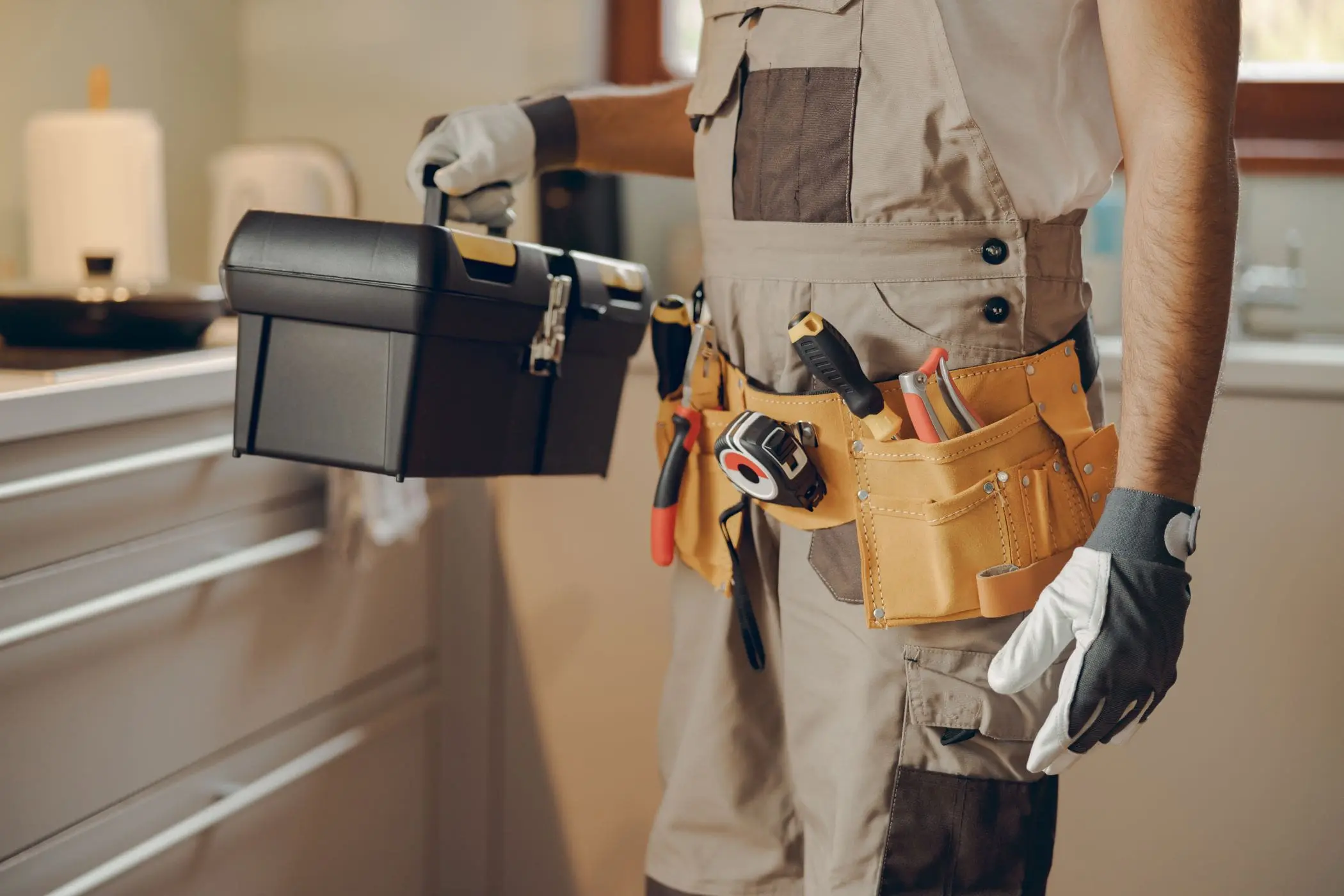 A close-up photograph of a construction worker's outfit and tools. The worker is wearing a beige and brown utility jumpsuit with multiple pockets and reinforced patches. A tan leather tool belt is secured around the waist, holding various tools including screwdrivers and pliers. The worker is wearing white work gloves and is holding a black toolbox in their left hand. The background is slightly blurred but shows a light-colored wall and a window. The image has a warm, professional quality with natural lighting. The composition is cropped to focus on the mid-section of the worker, showing the utility belt and tool arrangement in detail.