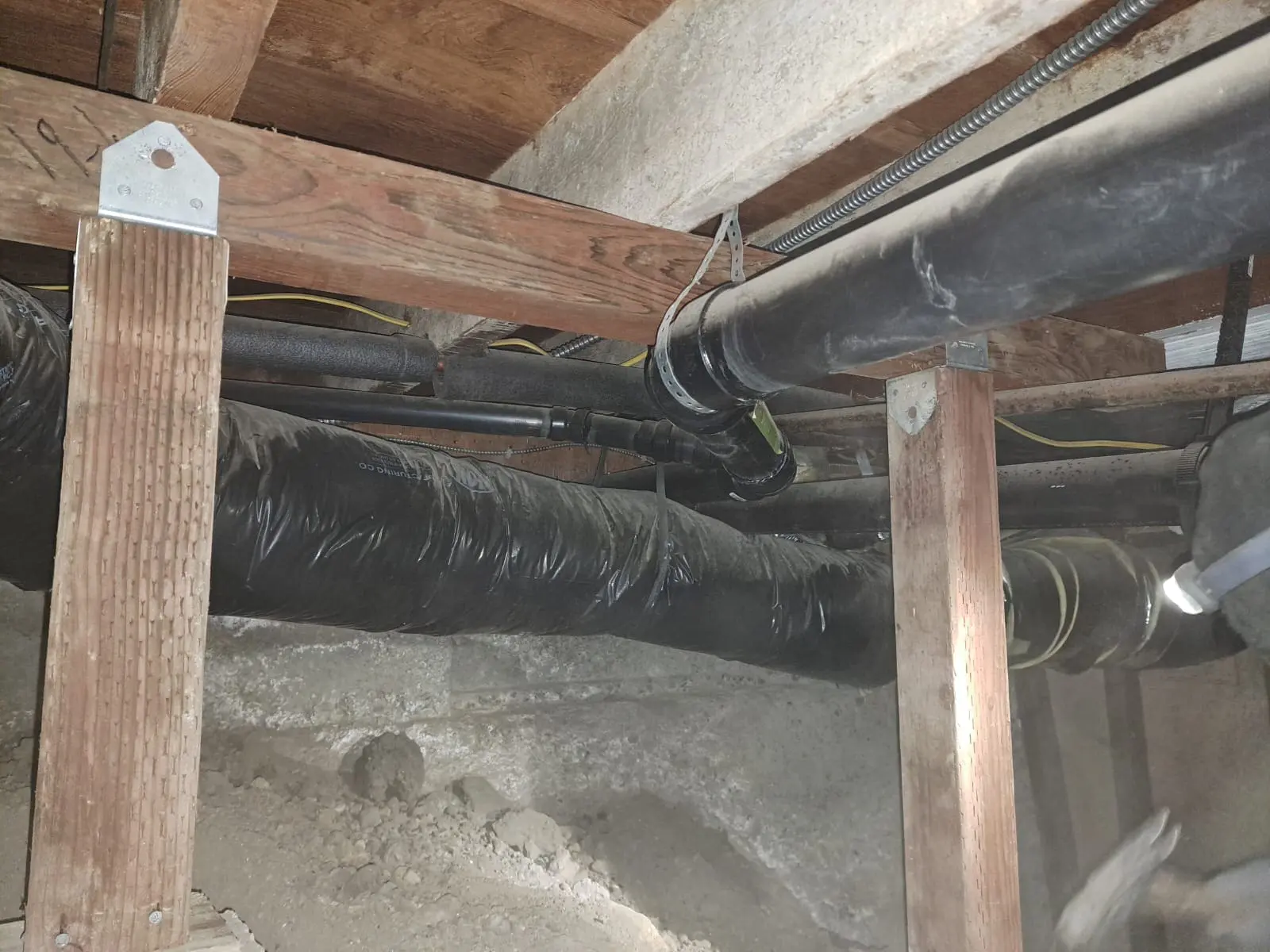 A close-up photograph of a basement crawlspace showing exposed wooden support beams and pipes. The wooden support beams are made of light-colored pine wood and are attached to a concrete foundation wall. Multiple black PVC pipes run horizontally across the frame, with one large insulated pipe wrapped in black rubber insulation. The pipes are connected with metal clamps and joints. The concrete foundation wall shows signs of age with a rough, weathered texture. The lighting is dim and creates shadows beneath the wooden beams. The image is taken from a low angle, looking upward at the structural elements. The concrete floor is visible at the bottom of the frame, showing a rough, unfinished surface with some debris or dirt scattered around.