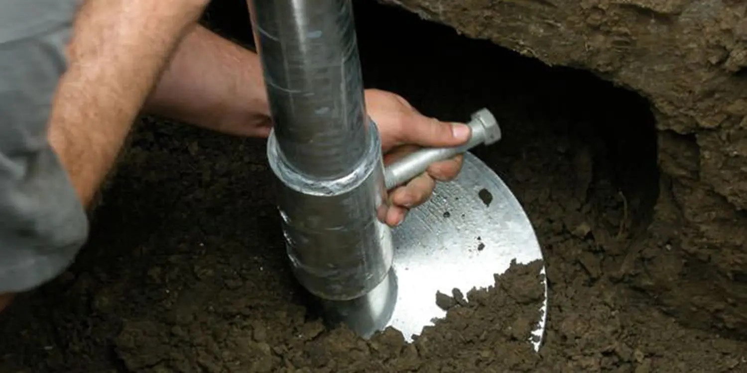 A close-up photograph of hands working with a metal auger in dark soil. The hands are gripping a large silver metal drilling tool with a cylindrical shape and ridged exterior. The tool is partially buried in rich, dark soil that fills the frame. The metal tool has a circular cutting edge visible at its tip, creating a contrast against the soil. The lighting creates reflections on the metallic surface of the tool, highlighting its industrial nature. The image is cropped to focus on the hands and tool, with the soil extending to the edges of the frame. The soil has a moist, freshly disturbed texture with small particles visible in the dark earth.