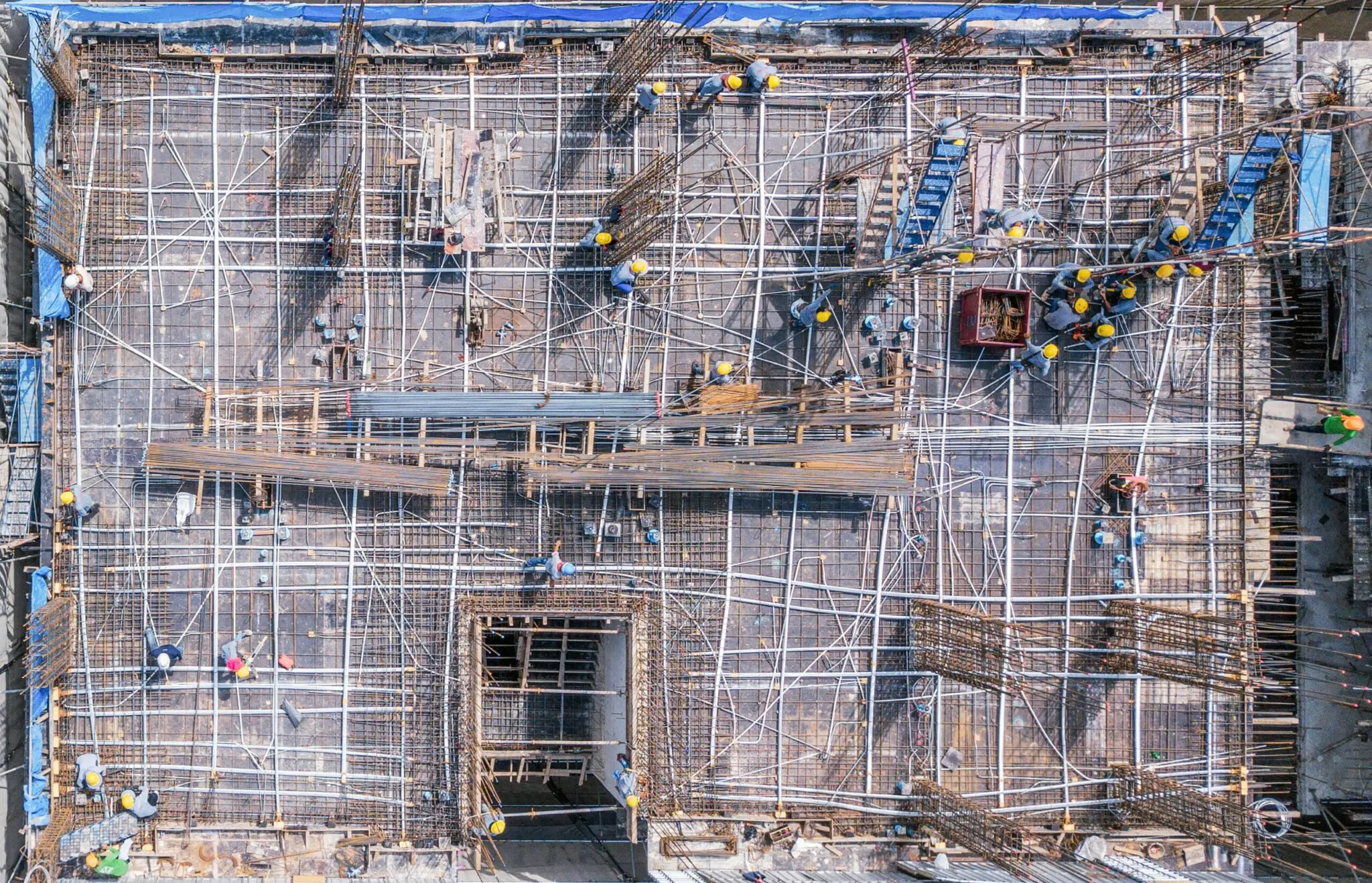 An aerial photograph of a construction site viewed from directly above. The image shows a large rectangular concrete structure under construction, with a complex network of metal scaffolding and support beams creating a geometric pattern across the entire surface. The scaffolding consists of silver-colored metal poles and crossbeams arranged in a grid pattern. Small yellow safety cones are scattered throughout the construction area. The concrete surface has a gray color and shows various construction openings and doorways. Blue tarpaulin is visible along the edges of the construction area. The image is taken from a high vantage point, showing the entire construction site in a top-down perspective. The lighting is even and natural, creating clear visibility of all construction elements. The construction site is a commercial or industrial building under development.
