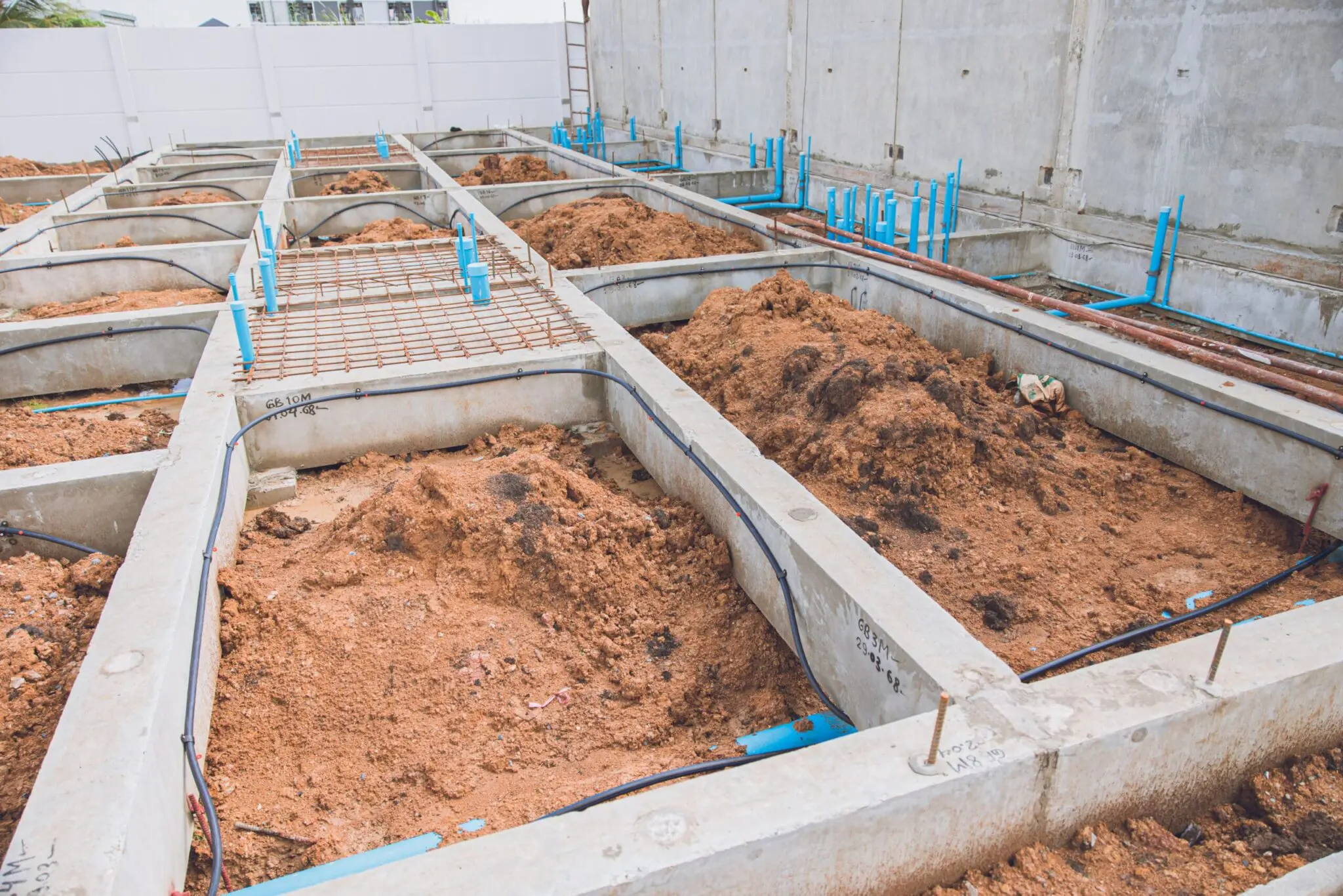 A construction site photograph showing exposed concrete foundation work in progress. The image shows multiple rectangular concrete foundation sections with reddish-brown soil and dirt filling the spaces between them. Blue PVC pipes run along the concrete borders, and small metal rebar grids are visible in one of the sections. The concrete foundation forms are light gray in color and have clean, straight edges. The background shows a white concrete wall. The soil is compacted and has a rich, reddish-brown color. The lighting is natural daylight, creating subtle shadows in the concrete forms. The image is taken from a high angle, showing the geometric pattern of the foundation layout.