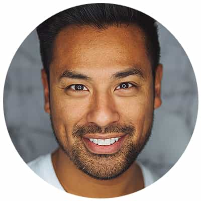 A professional headshot portrait in a circular frame. The subject is a middle-aged man with a warm, genuine smile and dark stubble beard. He has dark hair styled neatly to the side and bright brown eyes. He is wearing a white crew neck shirt. The background is a soft, blurred gray tone creating a professional studio atmosphere. The lighting is even and flattering, highlighting natural features and creating subtle shadows. The image has a clean, modern aesthetic with sharp focus on the facial features. The circular frame has a white border.