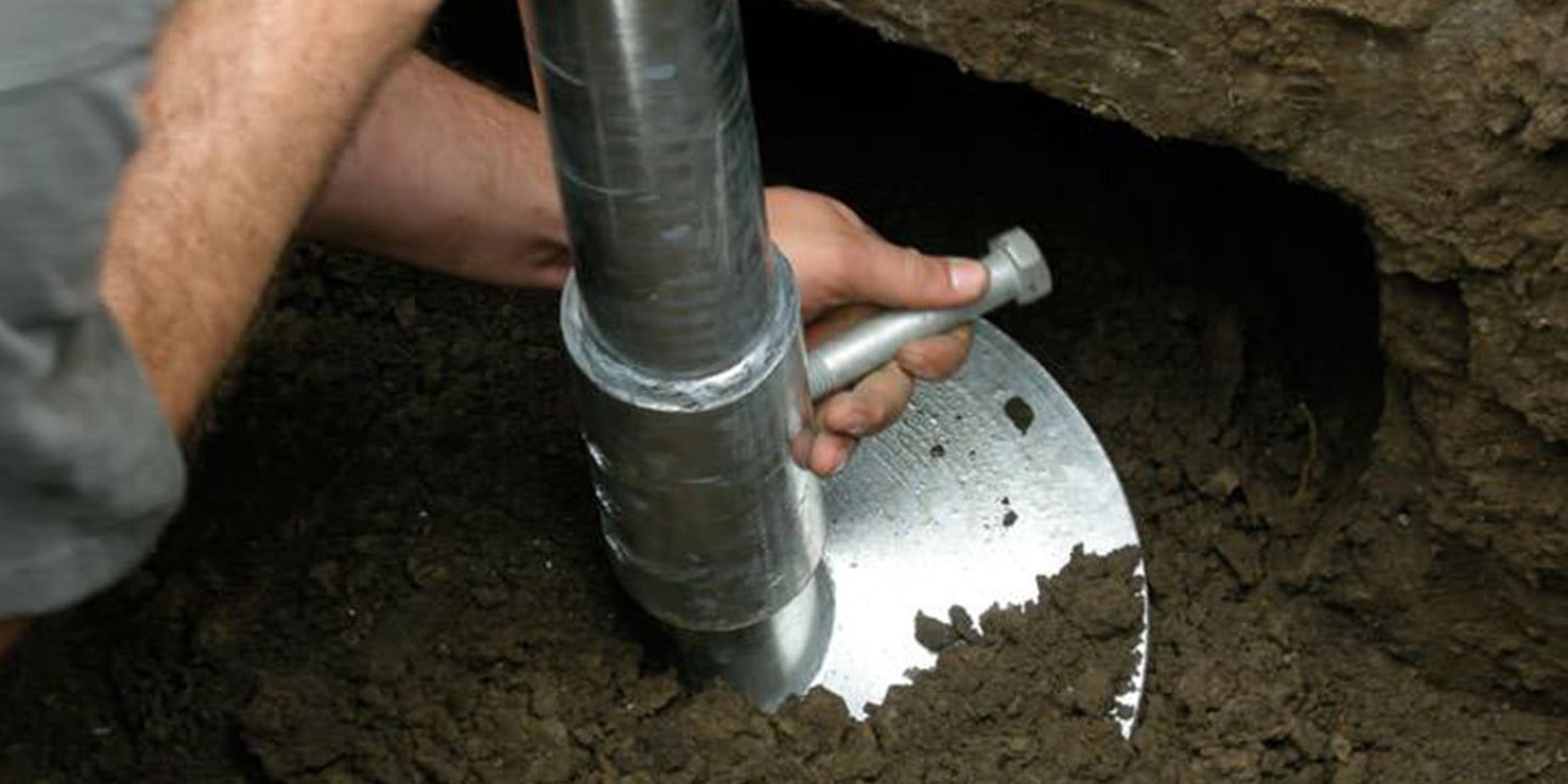 A close-up photograph of hands working with a metal auger in dark soil. The hands are gripping a large silver metal drilling tool with a cylindrical shape and ridged exterior. The tool is partially buried in rich, dark soil that fills the frame. The metal tool has a circular cutting edge visible at its tip, creating a contrast against the soil. The lighting creates reflections on the metallic surface of the tool, highlighting its industrial nature. The image is cropped to focus on the hands and tool, with the soil extending to the edges of the frame. The soil has a moist, freshly disturbed texture with small particles visible in the dark earth.