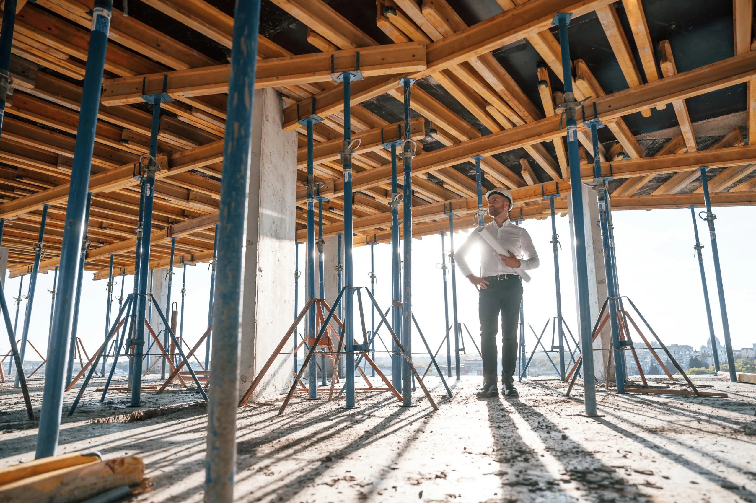 A professional architectural photograph of a construction site interior. A caucasian man in a crisp white dress shirt and dark pants stands beneath exposed wooden roof beams and metal scaffolding supports. The wooden beams are arranged in a geometric pattern, creating a diagonal framework across the ceiling. Blue metal scaffolding supports extend from floor to ceiling, creating vertical lines throughout the space. The floor is unfinished concrete with visible construction debris and dust. Natural sunlight streams in from the right side of the frame, casting dramatic shadows from the wooden beams onto the floor. The lighting creates a warm, golden glow on the wooden surfaces while maintaining cool tones in the metal elements. The image has a professional architectural photography style with sharp focus and high contrast. The composition is shot from a low angle, emphasizing the height and scale of the construction space.