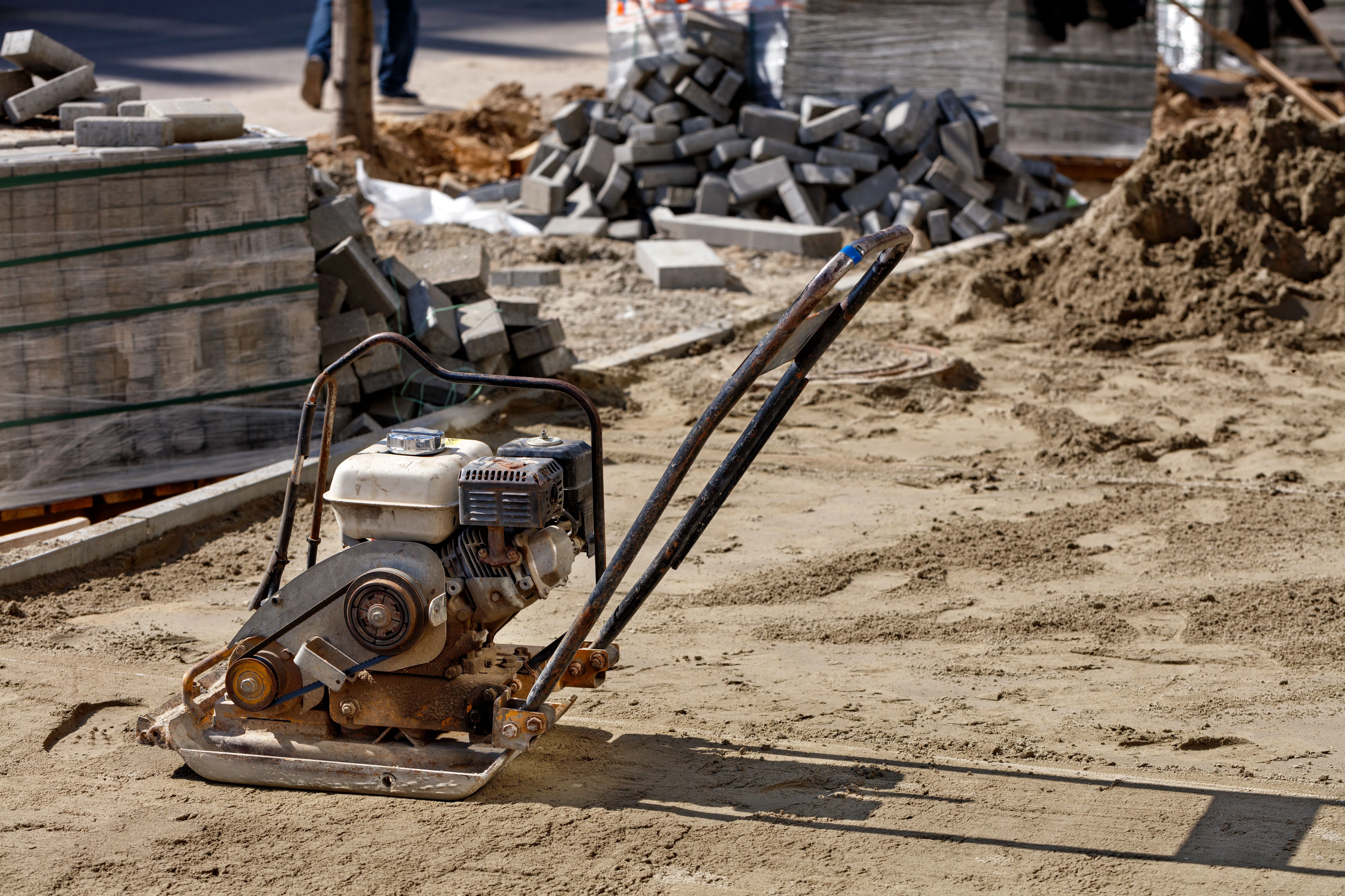 A close-up photograph of a construction-grade plate compactor on sandy soil. The compactor has a gray metal base plate with a black rubber handlebar and a white gas-powered engine with visible cooling fins and a pull-start cord. In the background, there is a pile of gray concrete blocks scattered on the ground. The soil is light beige in color and has a rough, granular texture. The image is shot from a low angle, with the compactor positioned diagonally across the frame. The lighting is natural and creates subtle shadows beneath the equipment. The background shows a construction site with partial views of other equipment and materials. The composition has a shallow depth of field, with the compactor in sharp focus while the background elements are slightly blurred.