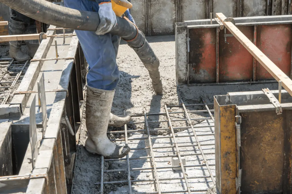A close-up photograph of construction work in progress. A worker wearing blue jeans, a light blue long-sleeved shirt, and gray rubber boots stands on metal rebar reinforcement bars. The worker is operating a gray concrete pump hose that extends out of frame. The scene shows a concrete foundation with metal formwork panels and wooden support structures. The formwork has a reddish-brown stain on its surface. The rebar creates a grid pattern across the concrete surface, with individual steel rods visible. The lighting is natural daylight, creating subtle shadows on the concrete surface. The image is cropped to focus on the worker's midsection and the construction materials, showing the industrial nature of the concrete pouring operation.