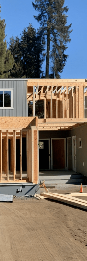 A construction site photograph of a two-story modern house with gray vertical siding in progress. The house features multiple white-framed windows and a wooden second-floor deck frame. The deck is supported by exposed wooden beams and posts. The foundation is visible with dirt and construction materials scattered around. The house has a contemporary architectural design with clean lines and a flat roof. In the background, tall evergreen trees, including Douglas firs, create a natural backdrop against a bright blue sky. The construction site shows exposed wooden framing for an attached garage on the left side of the house. Construction materials and orange safety cones are visible on the ground. The dirt around the foundation is freshly graded and is a light brown color. The image is taken from a slight angle, showing both the front and side of the house during the construction phase.